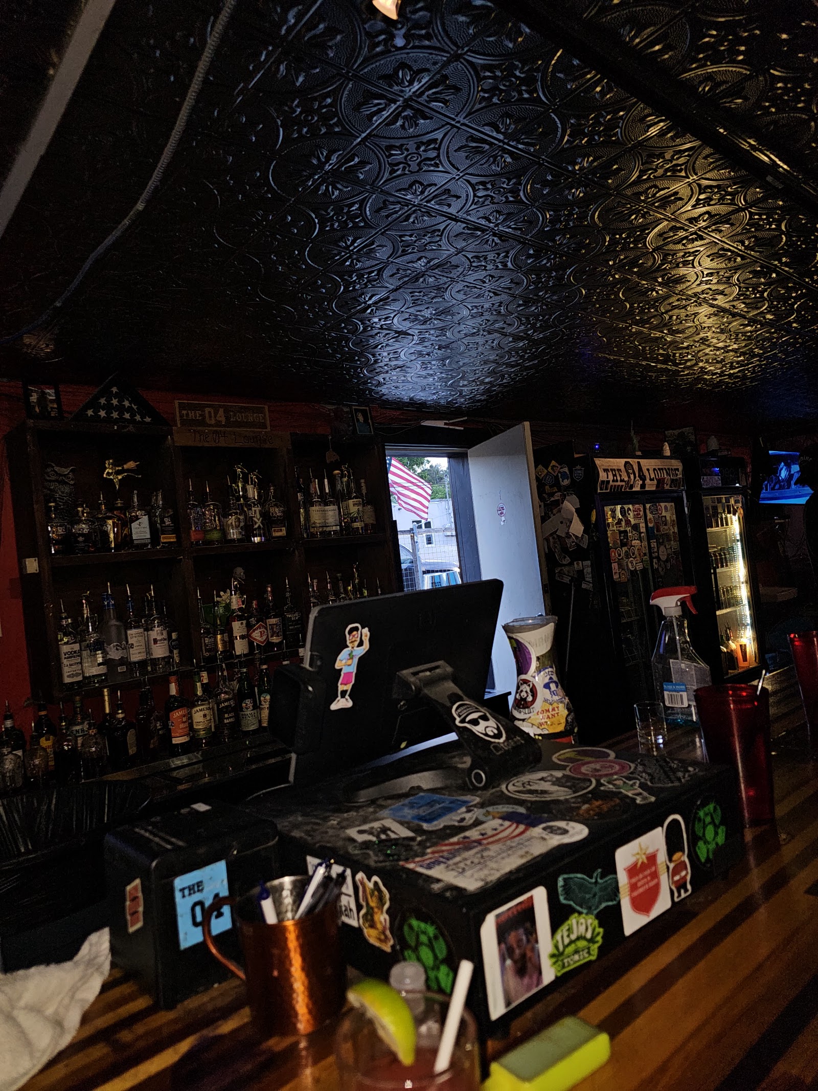 Behind the bar: sticker-covered register, open door to daylight, and the bottle shelves.